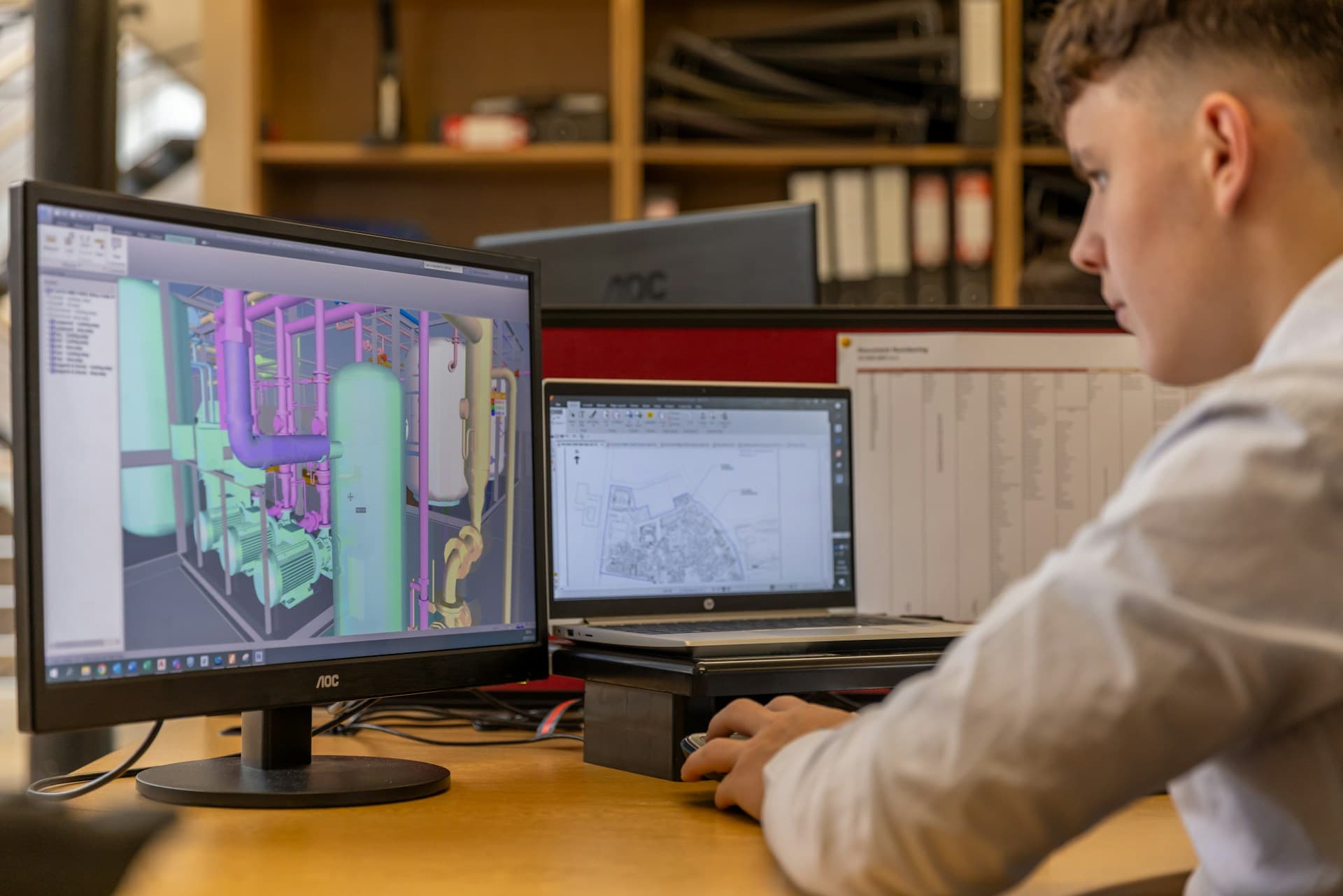 An engineer at a desk with a 3D model of pipework on the monitor and a 2D site plan open on a laptop